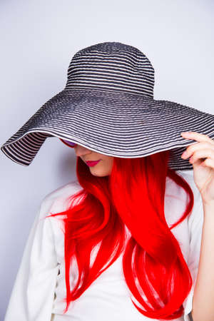 Attractive red-haired young woman in sunglasses in form of heart and striped white and black hat on white background. Sexy stylish model enjoying sun. Summertime.の写真素材