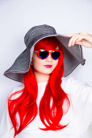 Attractive smiling red-haired young woman in sunglasses in form of heart and striped white and black hat on white background. Sexy stylish model enjoying sun. Summertime.の写真素材