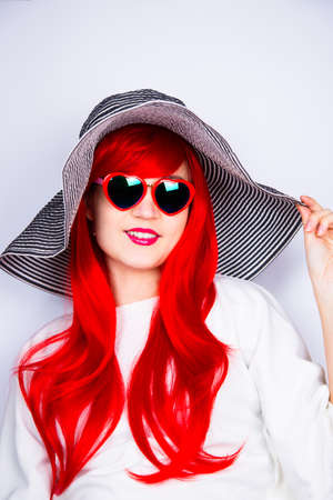 Attractive smiling red-haired young woman in sunglasses in form of heart and striped white and black hat on white background. Sexy stylish model enjoying sun. Summertime.の写真素材