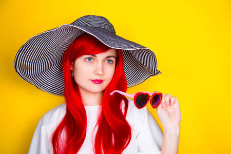 Attractive red-haired young woman in sunglasses in form of heart and striped white and black hat on yellow summer background. Sexy stylish model enjoying sun.の写真素材