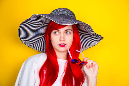 Attractive red-haired young woman in sunglasses in form of heart and striped white and black hat on yellow summer background. Sexy stylish model enjoying sun.の写真素材