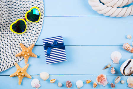 Hat and yellow sunglasses in shape of heart with shells, starfishes, gift box and sea rope on blue wooden background. Summer marine background. Beach accessories flat lay with negative space.の写真素材
