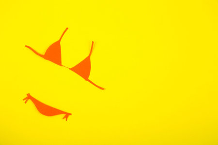 Orange paper bikini swimsuit on yellow background. Minimalistic summer colorful flat lay with negative space. Beach concept.の写真素材