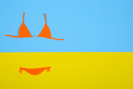 Orange paper bikini swimsuit on blue and yellow background. Minimalistic summer colorful flat lay with negative space. Beach concept.の写真素材