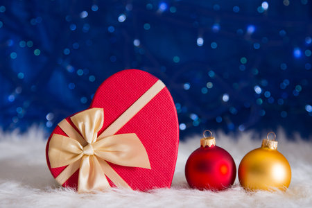 Red and golden Christmas balls with gift box in shape of heart on white fur with garland lights on blue bokeh background. New year card with empty space. Xmas festive decoration.の写真素材