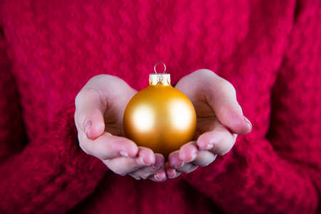 Beautiful golden Christmas ball in female hands on background of red knitted sweater. New year card. Xmas banner.の写真素材
