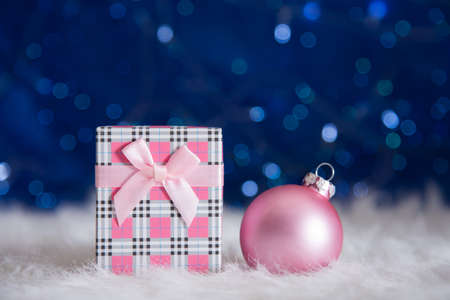 Pink gift box with beautiful Christmas ball on white fur with garland lights on blue bokeh background. New year card with empty space. Xmas festive decoration.の写真素材