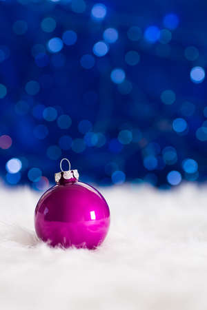 Crimson Christmas ball on white fur with garland lights on blue bokeh background. New year card with empty space. Xmas festive decoration.の写真素材