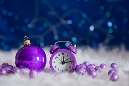 Purple little clock with Christmas ball and beads on white fur with garland lights on blue bokeh background.  New year card with empty space. Xmas festive decoration.の写真素材