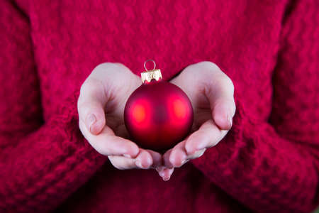 Beautiful red Christmas ball in female hands on background of red knitted sweater. New year card. Xmas banner.の写真素材