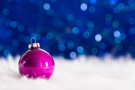 Crimson Christmas ball on white fur with garland lights on blue bokeh background. New year card with empty space. Xmas festive decoration.の写真素材