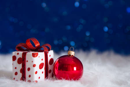 Beautiful gift box with red Christmas ball on white fur with garland lights on blue bokeh background. New year card with empty space. Xmas festive decoration.の写真素材