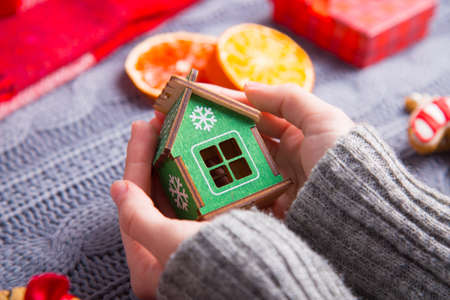 Female hands in gray knitted sweater holding green wooden house with Christmas decor on background. New Year decoration with dried citrus fruits, cookies, gifts and gingerbread. の写真素材