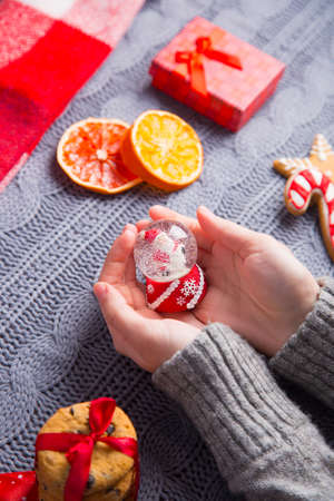 Female hands in gray knitted sweater holding glass snowball with Christmas decor on background. New Year decoration with dried citrus fruits, cookies, gifts and gingerbread. の写真素材