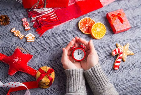 Female hands in gray knitted sweater holding little clock with Christmas decor on background. New Year decoration with dried citrus fruits, cookies, gifts and gingerbread. の写真素材