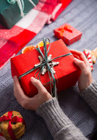 Female hands in gray knitted sweater holding red gift box with Christmas decor on background. New Year decoration with dried citrus fruits, cookies, gifts and gingerbread.の写真素材
