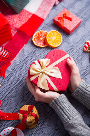 Female hands in gray knitted sweater holding gift box in shape of heart with Christmas decor on background. New Year decoration with dried citrus fruits, cookies, gifts and gingerbread.の写真素材