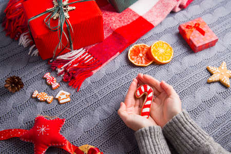 Female hands in gray knitted sweater holding glazed gingerbread in form of candy cane with Christmas decor on background. New Year decoration with dried citrus fruits, cookies, gifts and gingerbread.の写真素材