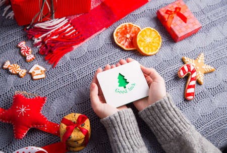 Female hands in gray knitted sweater holding greeting card with Christmas decor on background. New Year decoration with dried citrus fruits, cookies, gifts and gingerbread.の写真素材
