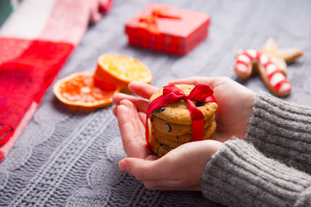 Female hands in gray knitted sweater holding homemade chocolate cookie in red ribbon with Christmas decor on background. New Year decoration with dried citrus fruits, cookies, gifts and gingerbread.の写真素材