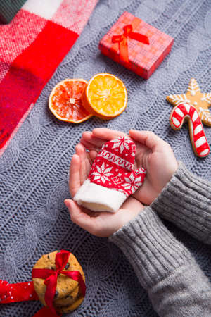 Female hands in gray knitted sweater holding little winter mitten with Christmas decor on background. New Year decoration with dried citrus fruits, cookies, gifts and gingerbread.の写真素材
