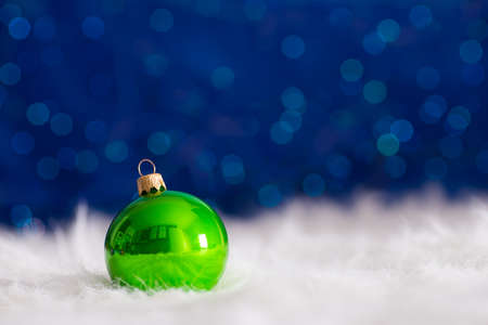 Green Christmas ball on white fur with garland lights on blue bokeh background. New year card with empty space. Xmas festive decoration.の写真素材