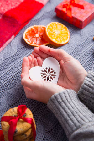 Female hands in gray knitted sweater holding white heart with Christmas decor on background. New Year decoration with dried citrus fruits, cookies, gifts and gingerbread.の写真素材