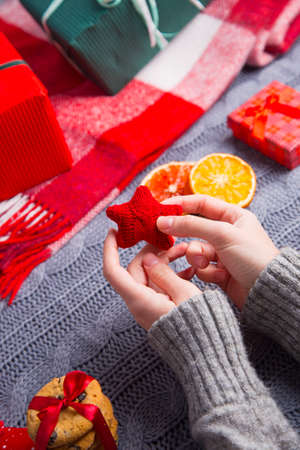 Female hands in gray sweater holding knitted red star with Christmas decor on background. New Year decoration with dried citrus fruits, cookies, gifts and gingerbread.の写真素材