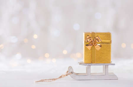 Little pretty golden gift box on toy white sled on light bokeh background. New year card. Xmas decoration.の写真素材