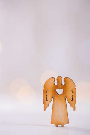 Wooden icon of Christmas angel with hole in form of heart on light bokeh background. Xmas card. Concept of love of religionの写真素材