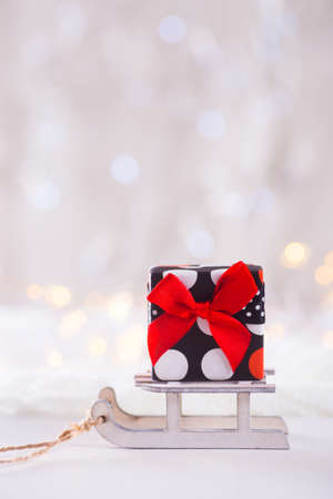 Little pretty spotted gift box with red bow on toy white sled on light bokeh background. New year card. Xmas decoration.の写真素材