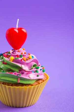 Closeup festive creamy cupcake with sprinkles and candle in form of red heart on purple background. Romantic cake for Valentineâs dayの写真素材