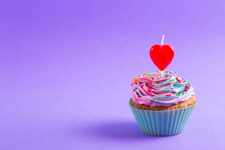 Festive creamy cupcake with sprinkles and candle in form of red heart on purple background. Romantic cake for Valentineâs dayの写真素材