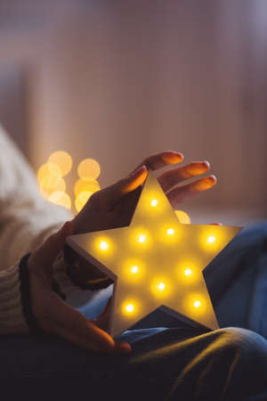 Young female hands holding glowing white LED star on warm bokeh background indoor. Festive Christmas illumination, holiday atmosphere.の写真素材