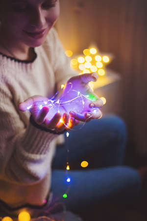 Pretty young woman in sweater holding warm glowing garland indoor. Preparation to Christmas party at home. の写真素材