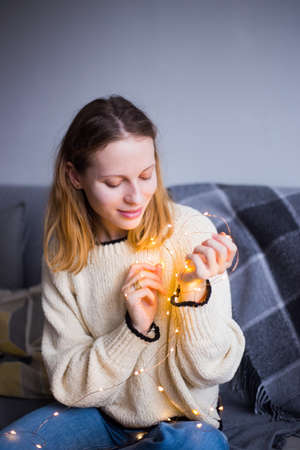 Pretty young woman in sweater holding warm glowing garland indoor. Preparation to Christmas party at home. の写真素材