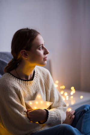 Pretty young woman in sweater holding warm glowing garland indoor. Preparation to Christmas party at home. の写真素材