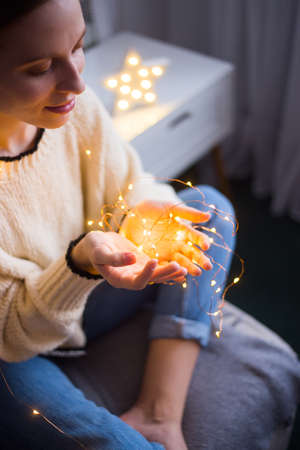 Pretty young woman in sweater holding warm glowing garland indoor. Preparation to Christmas party at home. の写真素材
