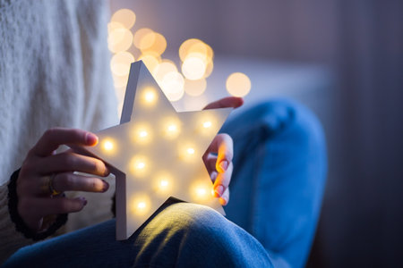 Young female hands holding glowing white LED star on warm bokeh background indoor. Festive Christmas...