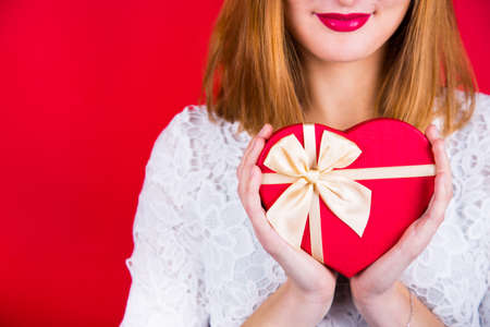 Young smiling woman holding red gift box in shape of heart on red background. Romantic surprise, St. Valentines Day present.の写真素材