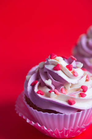 Close-up two cupcakes with creamy pink and white top decorated with little hearts on red background. St. Valentine dessert.の写真素材