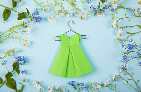 Green paper origami dress on hanger surrounded with blue and white little flowers on light mint background. Concept of spring fashion.の写真素材