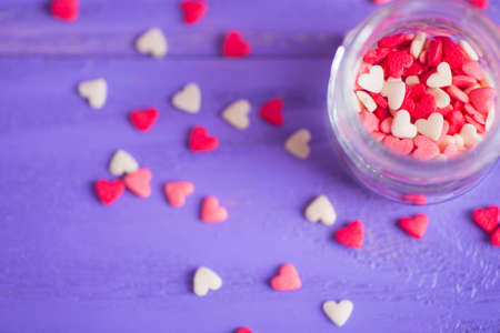 Open glass jar filled with many little candies in form of hearts on purple wooden background. St. Valentine Day card.の写真素材
