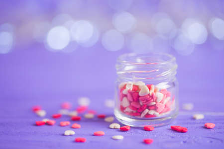 Open glass jar filled with many little candies in form of hearts on purple bokeh background. St. Valentine Day card.の写真素材