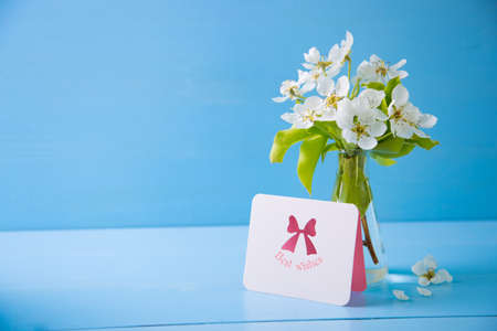 Greeting card with inscription "Best wishes" and bouquet of beautiful tender white flowering branches in glass vase on blue wooden background. Spring composition.の写真素材