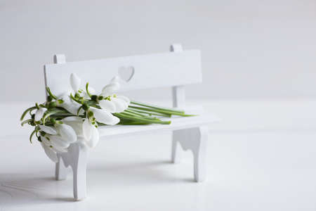 Bouquet of beautiful tender white snowdrops on white bench with hole in form of heart on light background. Spring composition.の写真素材