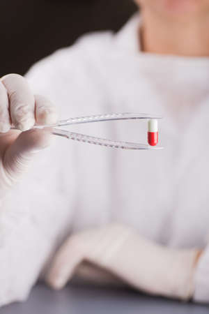 Hand in latex glove holding tweezers with red and white capsule. Study of medicine. Pharmaceutical research.の写真素材