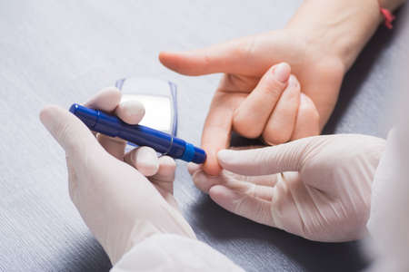 Doctor's hand in medical latex glove checking patient's blood glucose level using syringe pen. Glucometer on the table. Procedure for glucosometry.の写真素材