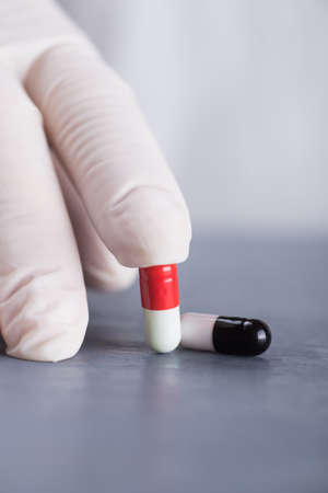 Closeup hand in medical latex glove holding two different capsules on gray table.の写真素材
