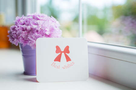Little purple bucket with tender bouquet of beautiful pink carnation and greeting card with inscription "Best wishes" near window in daylight. Spring motherâs day gift.の写真素材